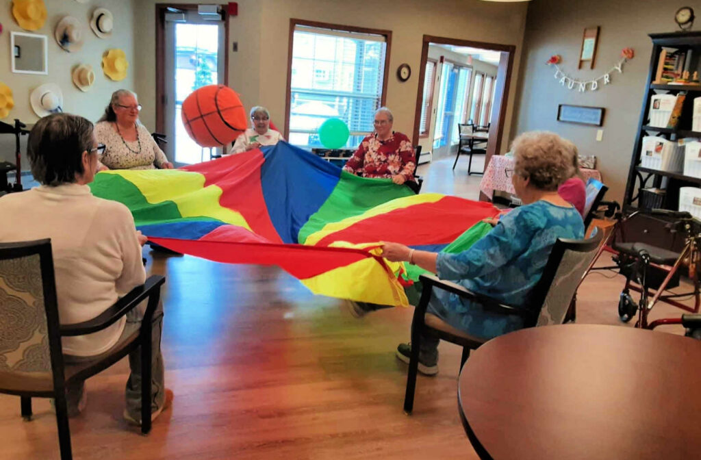 A group of seniors engaged in a fun activity that involves bouncing around a stuffed big ball on a colourful nylon sheet.