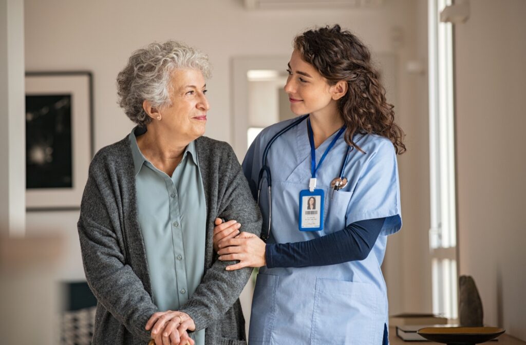 A caregiver in a blue uniform supports an older adult as they walk together indoors