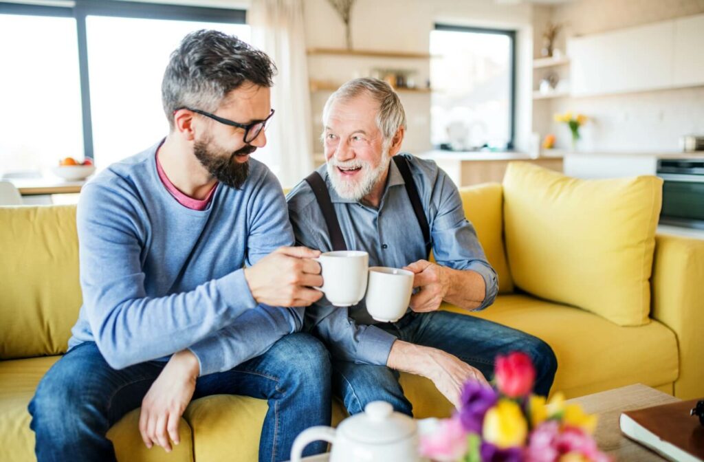 A senior and their adult child sitting on a yellow couch in a bright living room sharing coffee